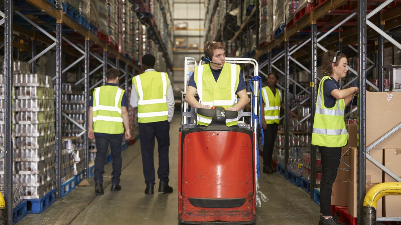 Woman selects box in warehouse for coworker on tow tractor